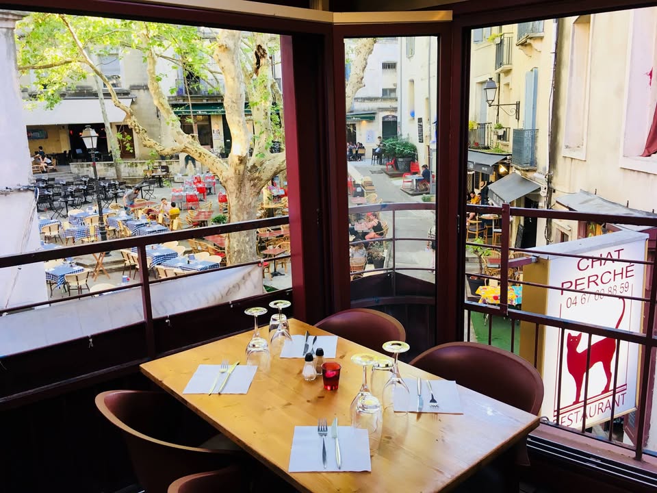 Table dressée en bois clair devant les baies vitrées de la salle du haut ; en dessous, la place de la Chapelle Neuve, son platane, les chaises rouges et les nappes à carreaux de la terrasse.
