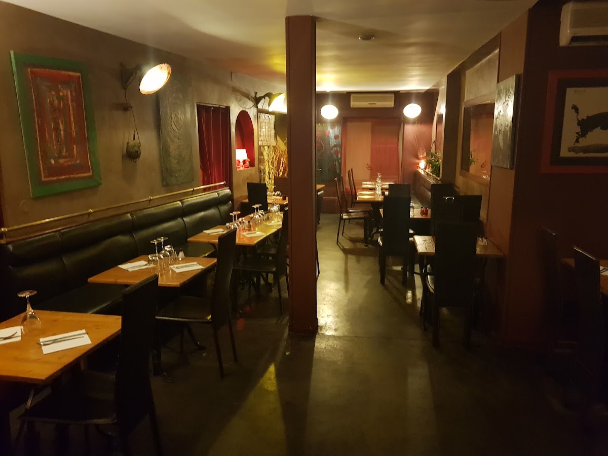 La salle du bas : longue banquette en cuir sombre le long du mur, barre de laiton au-dessus, tableaux d'art populaire accrochés serré, colonne bordeaux au centre et alcôve à rideau rouge au fond.