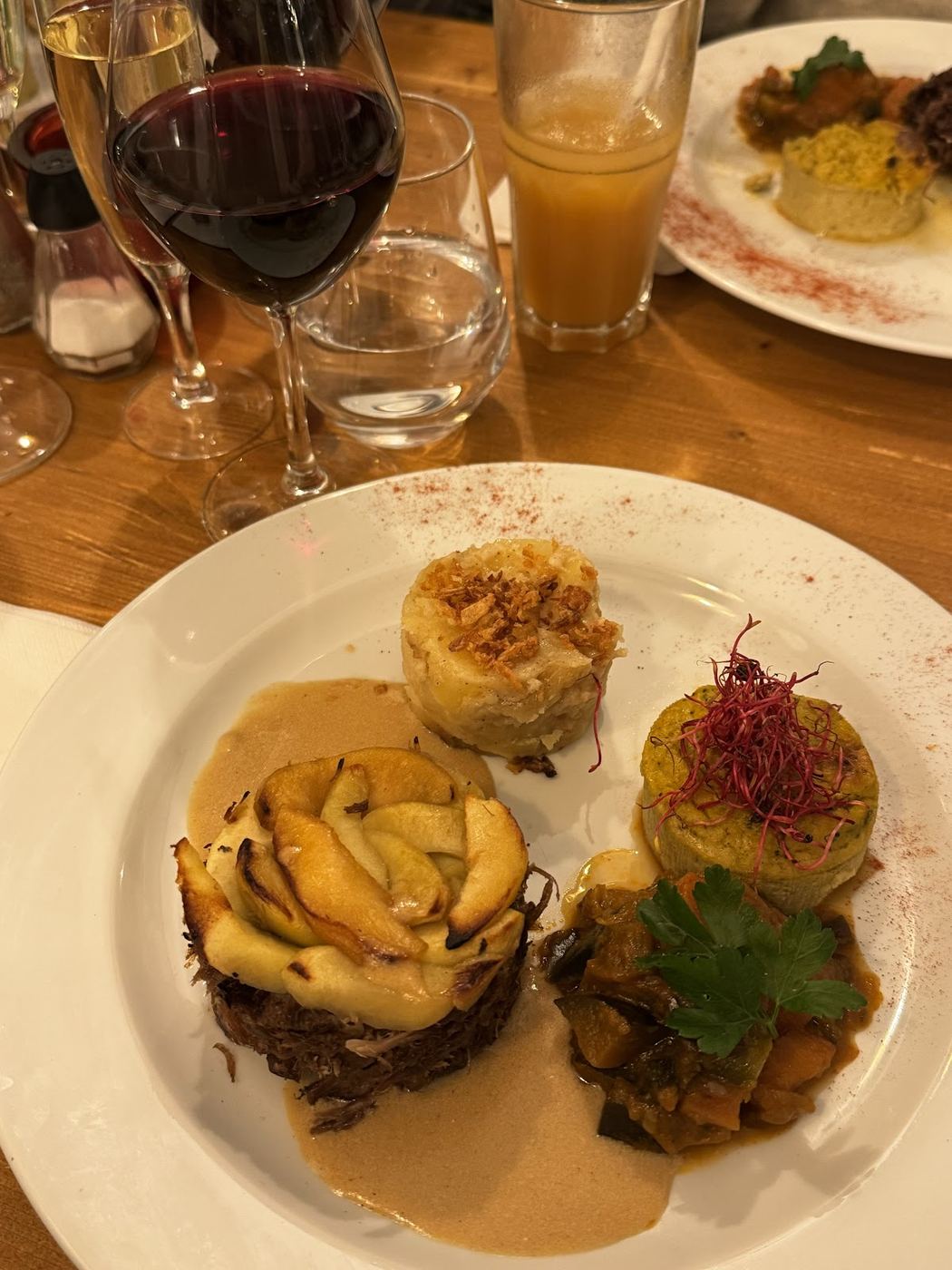 Assiette servie en salle : viande confite effilochée sous des lamelles dorées, gratin, flan de courgette et ratatouille, avec un verre de vin rouge sur la table en pin.