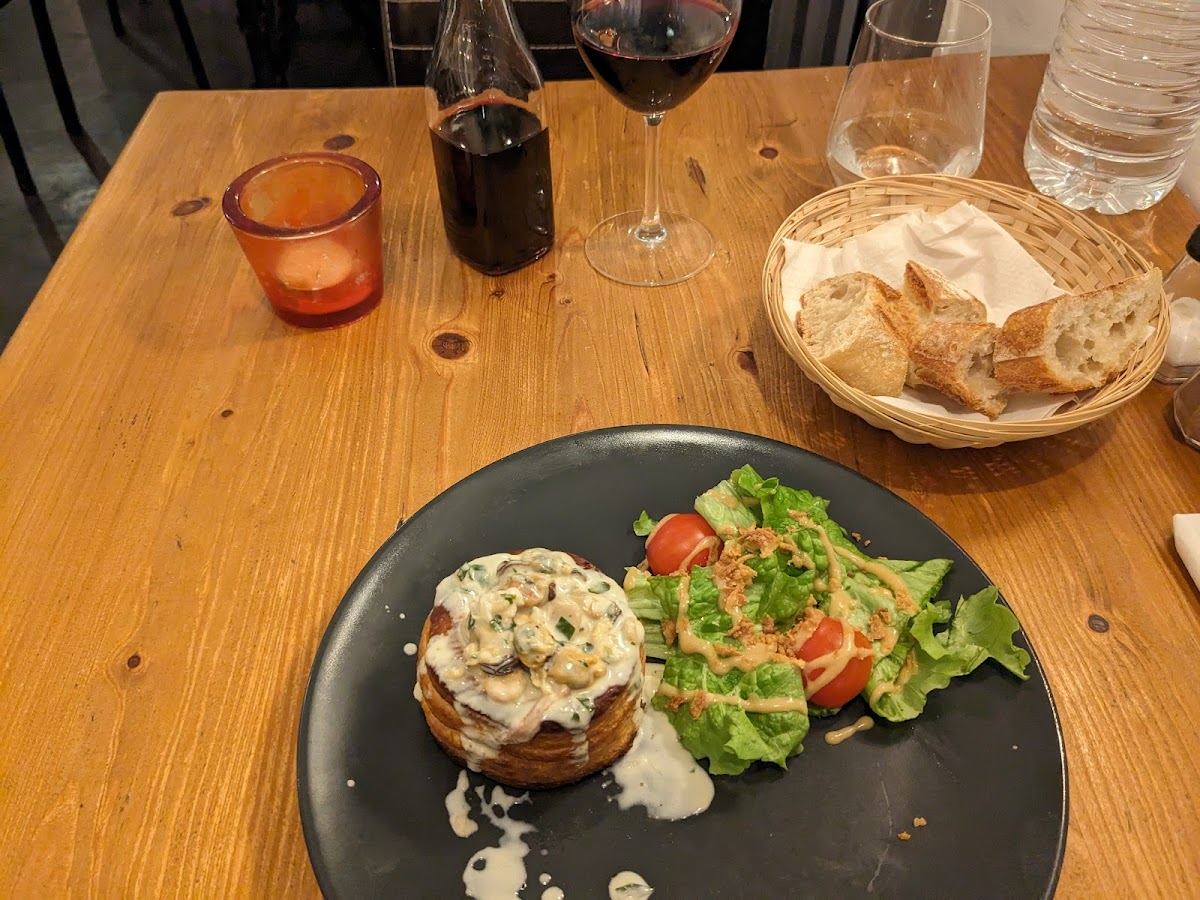 Une table en pin avec un feuilleté nappé de sauce aux champignons, une petite salade, une corbeille de pain et une carafe de vin rouge.